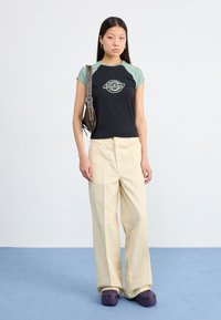 A young person stands on blue carpeting, wearing a black and green graphic t-shirt, beige pants, and purple clogs, holding a brown bag.