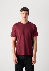 Een man staat recht vooruit met een effen bordeauxrode T-shirt met korte mouwen en zwarte broek tegen een effen lichte achtergrond.