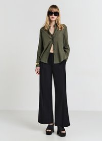 Camicia verde oliva con bottoni, finitura strutturata, abbinata a pantaloni neri larghi. Il modello indossa occhiali da sole neri oversize e scarpe con zeppa.