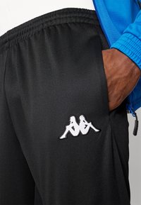 Pantalon de sport noir en tissu lisse avec une taille élastique. Présente un logo Kappa brodé en blanc sur le côté gauche.