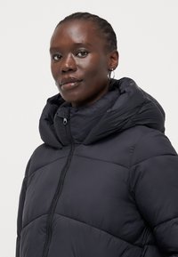 Svart pufferjakke med høy krage, synlige sømmer, glidelås foran og en glatt, blank tekstur. Har hette med rynkedetalj.