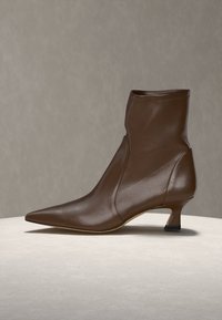 Bottine en cuir marron avec bout pointu et petit talon kitten sur une surface claire devant un fond uni.