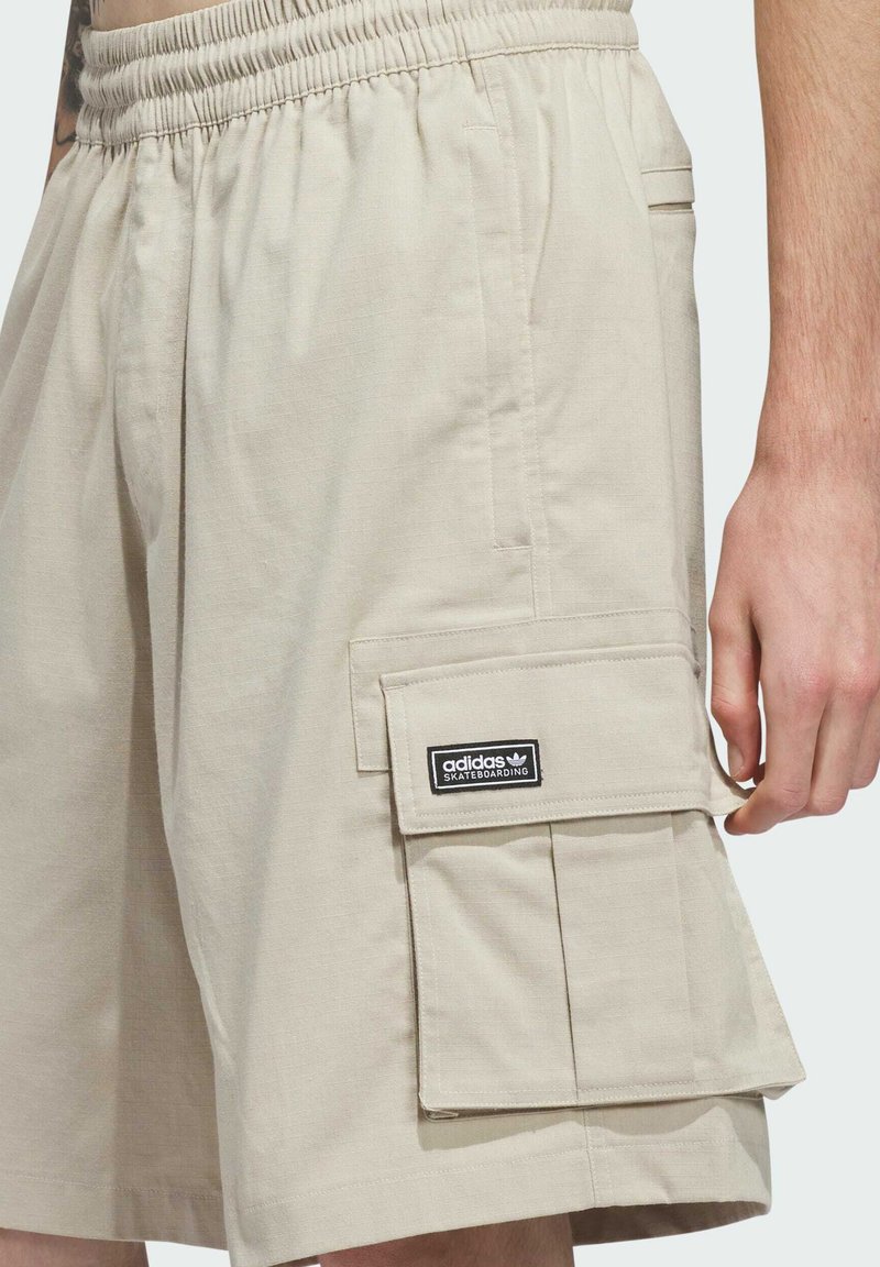 Pantalones cortos cargo beige con cintura elástica y solapa lateral del bolsillo etiquetada "adidas Skateboarding" usados por una persona con brazo visible.