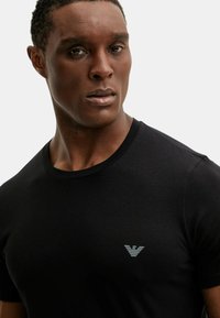 T-shirt nera in cotone con scollo girocollo, con un piccolo logo grigio chiaro sul lato sinistro del petto. Tessuto liscio, vestibilità standard.