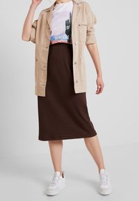 Beige übergroße Jacke, weißes T-Shirt mit Grafiken einer Wüstenlandschaft, brauner gerippter Midirock und weiße Sneakers mit strukturierten Sohlen.