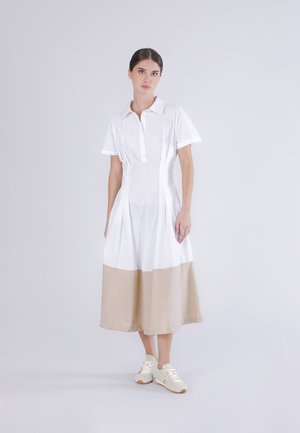 Donna in piedi che indossa un vestito bianco con maniche corte, collo e orlo beige, abbinato a scarpe da ginnastica bianche, su uno sfondo semplice.