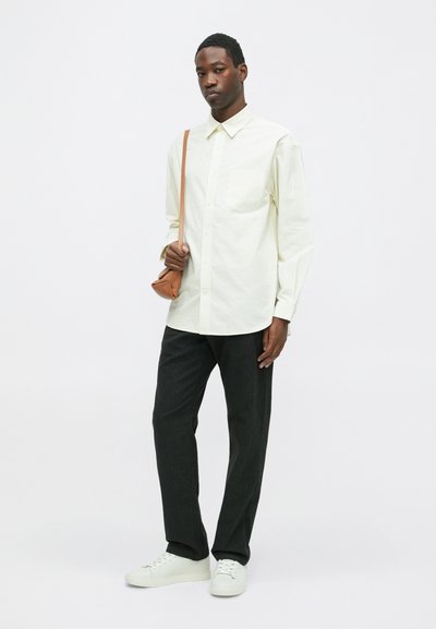 Jasno beżowa koszula zapinana na guziki z kieszenią na piersi, połączona z ciemnymi spodniami i białymi trampkami. Brunatna torba na ramię dopełnia wygląd.