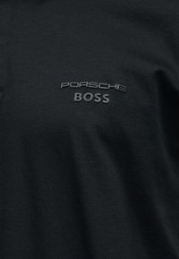 Μαύρο ύφασμα με ανάγλυφο κείμενο "PORSCHE" πάνω από το "BOSS" στο κέντρο του στήθους, που δείχνει λεπτή υφή και σκιές.