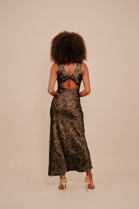 Göndör hajú nő egy ujjatlan, sötét virágmintás ruhában, hátsó kivágással és arany magassarkú szandálban áll egy egyszínű háttér előtt.