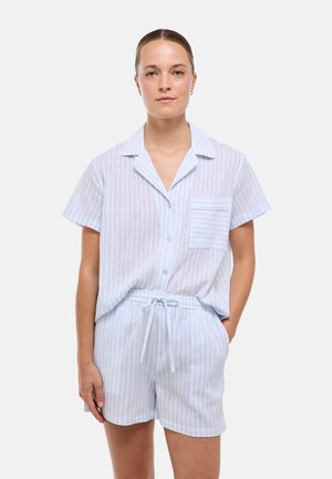 Femme portant une chemise boutonnée à manches courtes rayée bleu clair et blanc avec un short à cordon assorti, debout avec une main dans la poche.