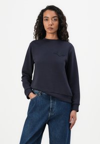 Kvinne med krøllete hår iført en marineblå genser med en liten brodert logo og løse blå jeans, stående foran en hvit bakgrunn.