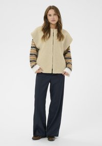 Bež prsluk od flisa s kap rukavima i prugastim pletenim rukavima u nijansama smeđe i krem, u kombinaciji s tamnoplavim širokim hlačama.