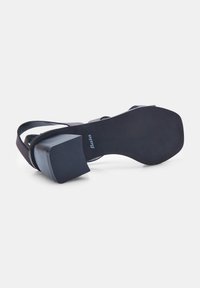 Crne sandale na visoku petu s teksturiranim gumenim potplatom, kutnim blok-petama i širokim trakama. Vidljivo brendiranje na potplatu.