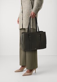 Sac en cuir texturé vert avec deux longues poignées, accents dorés et fermeture éclair, associé à un pantalon beige fuselé et des talons.