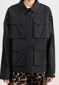 Svart jacka med stora fickor fram, krage och långa ärmar. Tillverkad av slätt tyg med knäppning framtill.