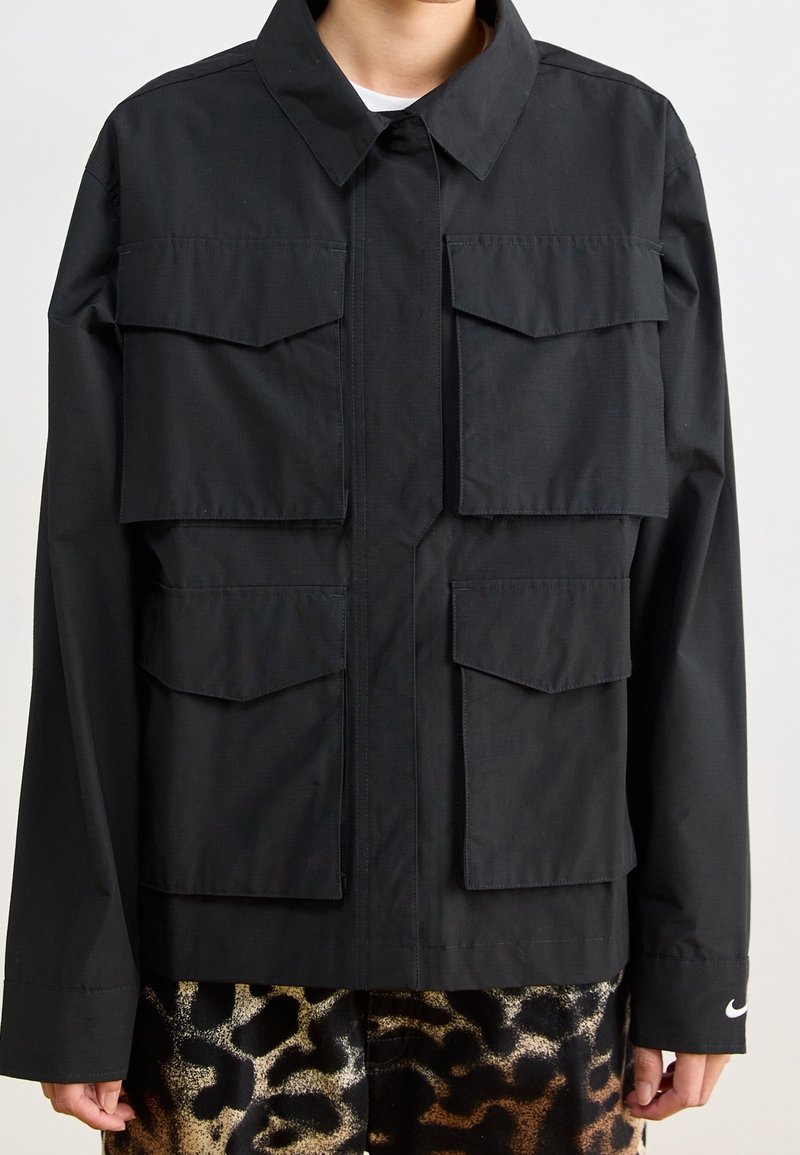 Svart jacka med stora fickor fram, krage och långa ärmar. Tillverkad av slätt tyg med knäppning framtill.