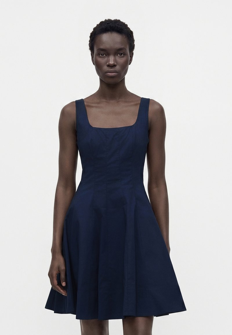 Femme aux cheveux courts et bouclés portant une robe bleu marine sans manches avec un décolleté carré, debout devant un fond blanc uni.
