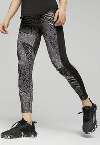 Person, der schwarze und graue Tierdruck-Leggings und schwarze Sportschuhe trägt, steht mit gespreizten Beinen vor einem neutralen Hintergrund.