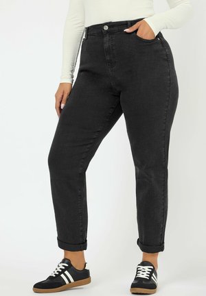 Kvinde iført sort høj talje-jeans med opklappede kanter, hvid langærmet top og sorte sneakers med hvide striber, stående med den ene hånd i lommen.