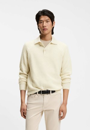 Jonge man met zwart haar die een crèmekleurige, getextureerde trui met lange mouwen en lichtbeige broek met een zwarte riem draagt, staand tegen een effen achtergrond.
