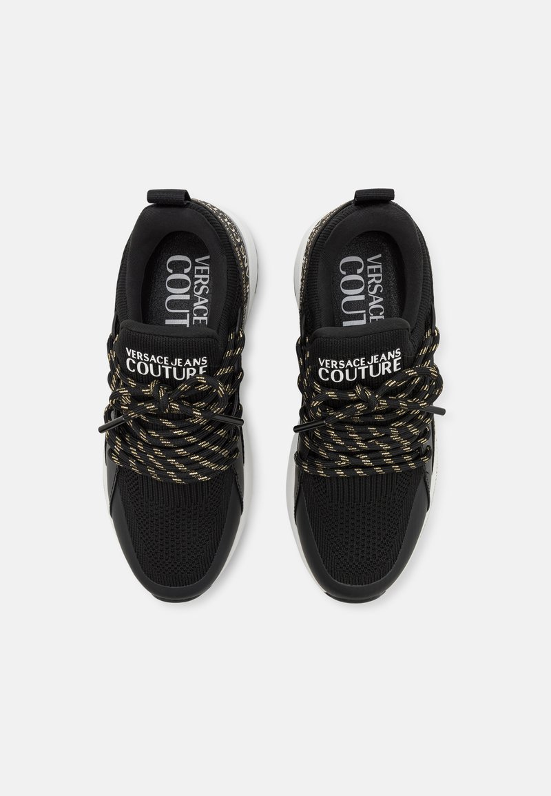 Czarne sneakersy z teksturowanym, dzianinowym wierzchem, refleksyjnymi akcentami i złotymi sznurowadłami w wzory. Posiadają wyściełany język i szczegóły logo po bokach.