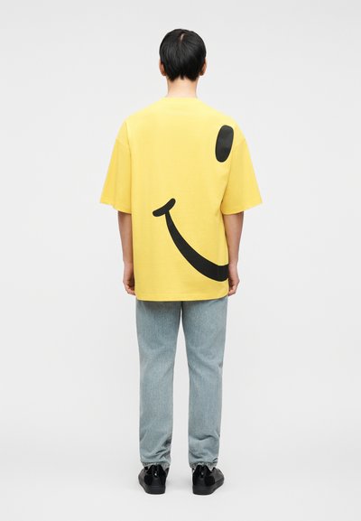 Κίτρινο βαμβακερό μπλουζάκι με μεγάλο μαύρο γραφικό χαμογελαστό πρόσωπο στην πλάτη. Το μοντέλο φοράει ανοιχτό μπλε τζιν και μαύρα παπούτσια, εμφανίζεται από πίσω.