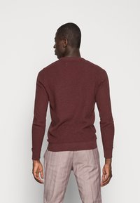 Burgunderfarbener Pullover mit langen Ärmeln und Rundhalsausschnitt. Strukturierter Stoff und gerippter Saum. Getragen mit hellen karierten Hosen.