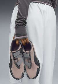 Rosa atmungsaktive Sneakers mit grauen Riemen und schwarzen Akzenten, strukturierten Sohlen und runder Form, von einer Hand vor einem schlichten Hintergrund gehalten.