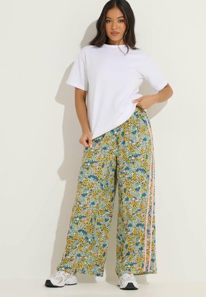 Frau mit langen dunklen Haaren, die ein schlichtes weißes T-Shirt und weite geblümte Hosen mit rosa Streifen trägt, steht vor einem hellen Hintergrund.