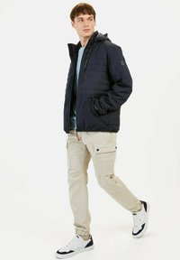 Navy zip-up jacket with a hood, featuring a quilted design, paired with light beige cargo pants and white sneakers with navy accents.