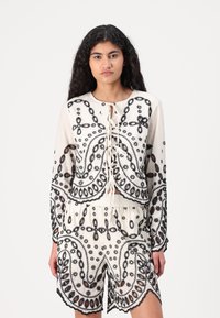 White embroidered two-piece set featuring long-sleeve top with tie closure and drawstring shorts, detailed with black floral patterns.