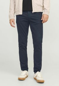 Dunkelblaue Chinos aus Baumwolle, mit Slim Fit und seitlichen Taschen, kombiniert mit weißen Sneakers und einer leichten Jacke. Einfaches, klares Design.