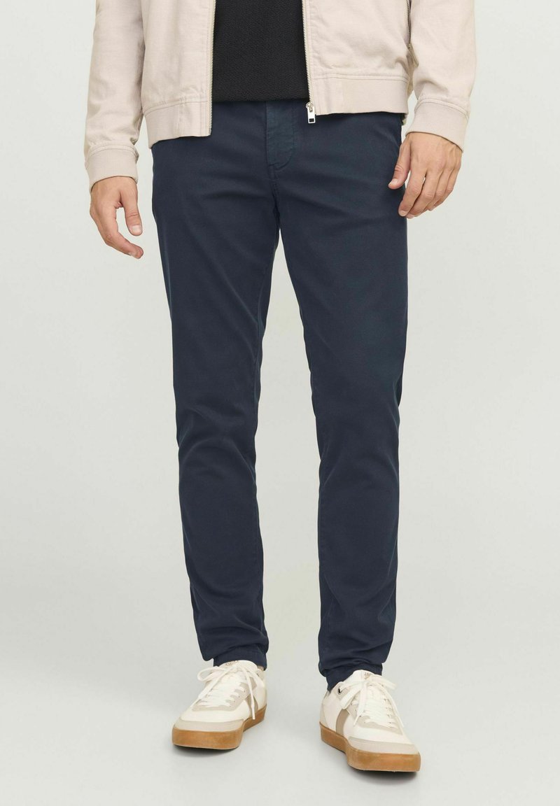 Dunkelblaue Chinos aus Baumwolle, mit Slim Fit und seitlichen Taschen, kombiniert mit weißen Sneakers und einer leichten Jacke. Einfaches, klares Design.