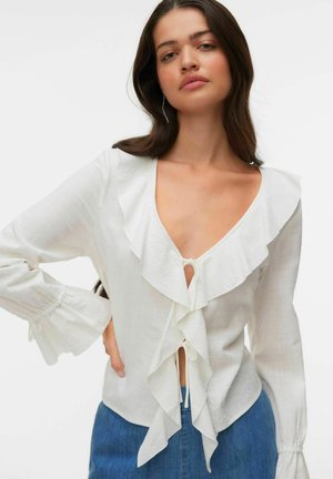 Nő fehér, hosszú ujjú blúzt visel fodros mély V-nyakkivágással és elöl megkötővel, kék farmerral, sima fehér háttér előtt.