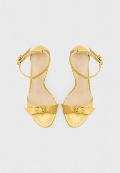 Χρυσά γυναικεία παπούτσια με υφή, ανοιχτά στο δάχτυλο, με λουράκι στον αστράγαλο και λεπτομέρεια κόμπου στο μπροστινό μέρος, παρουσιασμένα από πάνω σε ουδέτερο φόντο.