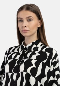 Schwarze und weiße gemusterte Bluse mit einem kragenförmigen Ausschnitt, glattem Stoff und abstrakten Formen, die ein wellenartiges Design auf der Oberfläche erzeugen.