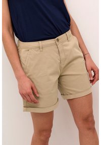 Short beige retroussé porté avec une chemise bleu marine rentrée, montrant des poches avant et une fermeture à boutons sur le short.