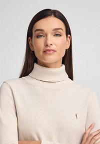 Maglione a dolcevita beige realizzato in morbido tessuto, con un design aderente e polsini a costine, caratterizzato da un discreto logo impresso sul petto.
