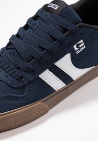Dark blue suede sneaker with white accents and stripes, featuring a round toe, textured laces, and a brown rubber outsole.