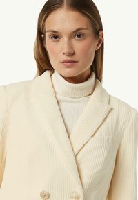 Cremfarbener gerippter Blazer mit Doppelknöpfen und Revers, über einem passenden gerippten Rollkragenpullover geschichtet. Subtile Details des strukturierten Stoffes.