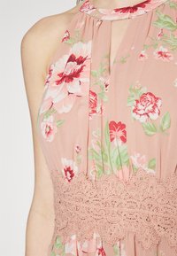Kvetinové šaty s ružovým pozadím, so vzormi červených a zelených kvetov. Čipkový detail v oblasti pásu, halterneckový strih s záhybmi.