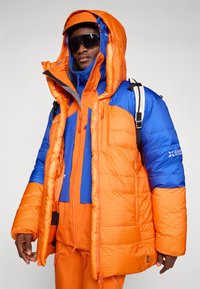 Chaqueta tipo puff en naranja y azul, con una gran capucha, textura de patrón en cuadrícula, puños ajustables y bolsillos con cremallera. Acompañada de gafas de sol.