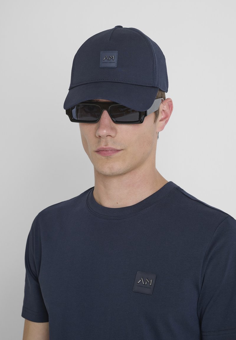 Cappellino da baseball blu scuro con una toppa logo sul davanti, indossato con occhiali da sole neri a forma quadrata e una maglietta blu scura abbinata con una piccola toppa logo.