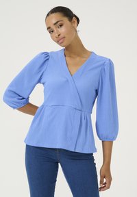 Top avvolgente azzurro chiaro, con texture, maniche a tre quarti a sbuffo e vita a peplum, accentuato da un profondo scollo a V e tessuto morbido.
