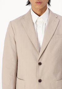 Beige textured blazer with a notched lapel, featuring two brown buttons, side pockets, and a white collared shirt underneath.