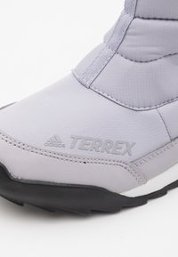 Grå syntetisk støvle med en tekstureret overflade, der har "TERREX"-logoet og en sort gummisål for greb. Vertikale sømme bemærket.