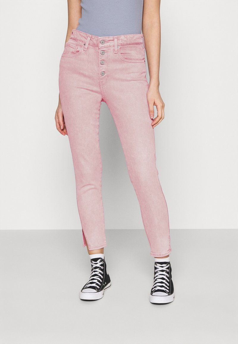 Personne portant un jean rose taille haute avec braguette à boutons et fentes latérales, associé à des baskets montantes noires et des chaussettes blanches.