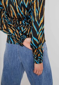 Camicia nera con motivo zigzag blu e oro, caratterizzata da dettagli elasticizzati in vita e polsini con bottoni, abbinata a jeans azzurri chiari.