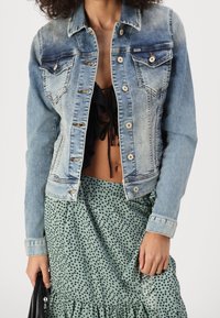 Chaqueta de denim azul claro con un diseño ajustado, bolsillos en el pecho y cierre de botones. Llevada sobre una blusa negra con volantes y una falda verde con puntos.