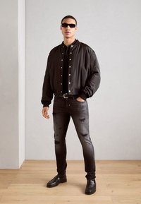 Chaqueta bomber negra con botones de presión plateados, camisa negra, jeans oscuros desgastados y mocasines negros. Modelo lleva gafas de sol negras.
